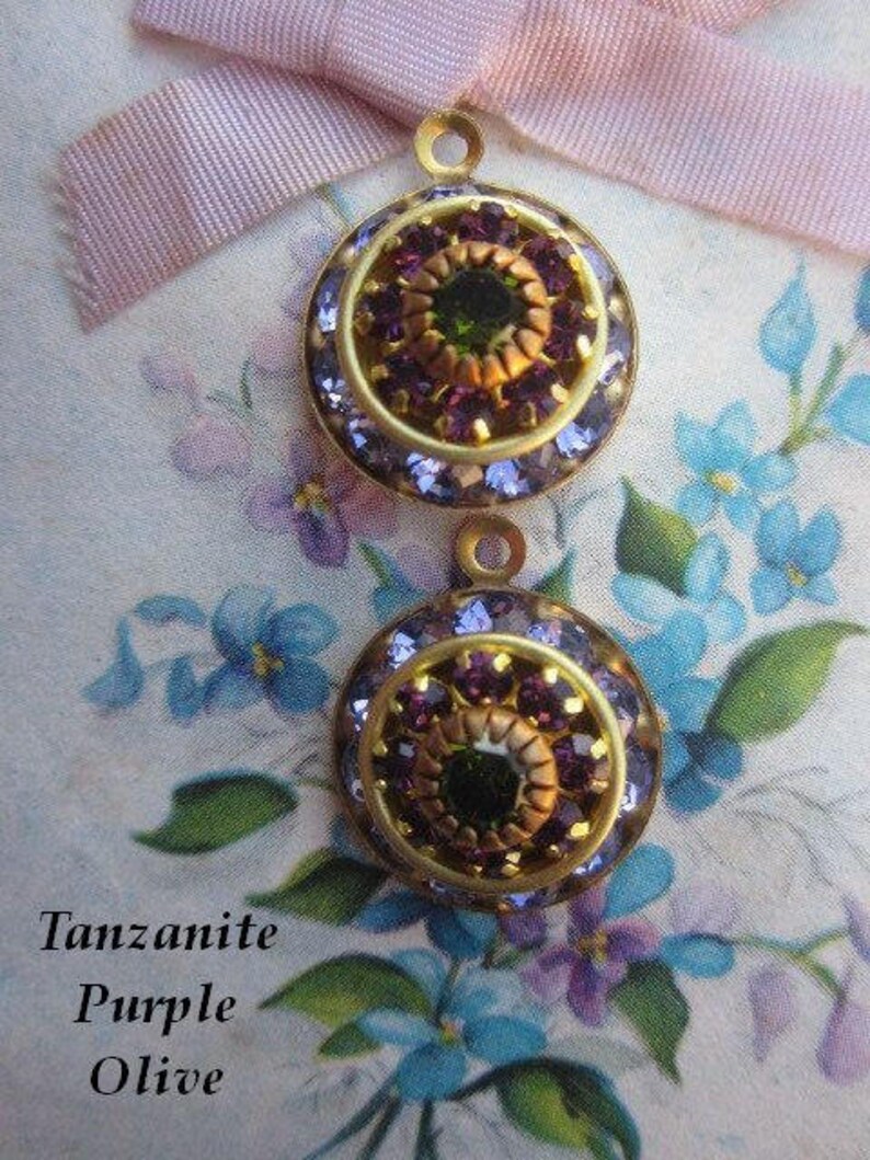 May include: Two gold-toned round pendants with a center cluster of purple and green rhinestones. The pendants have a textured surface and are surrounded by a ring of smaller purple rhinestones.