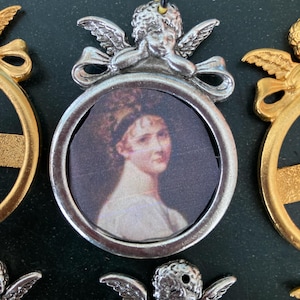 Peut inclure: Une décoration argentée en forme de chérubin tenant un petit cadre ovale avec un portrait de femme à l'intérieur. La décoration est ornée d'un nœud et d'une ficelle pour l'accrocher.