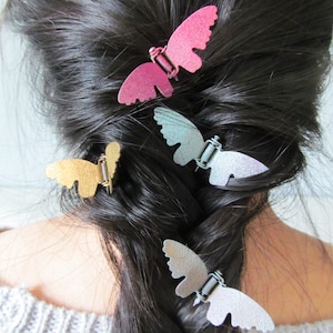 May include: A close-up of dark hair styled in a braid, adorned with several butterfly hair clips. The clips are in various metallic colors, including pink, silver, and gold. The butterfly clips are attached with a spring mechanism.