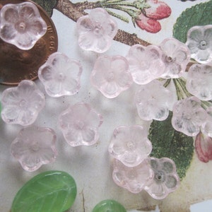 May include: A collection of pale pink glass flower beads with a clear center. The beads are arranged on a white surface with green leaves in the background.