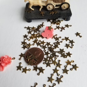 K&ouml;nnte beinhalten: Ein Haufen goldener sternf&ouml;rmiger Charms mit Sprungringen. Die Charms sind auf einer wei&szlig;en Oberfl&auml;che mit einem Penny als Gr&ouml;&szlig;envergleich verstreut. Im Hintergrund befinden sich eine rosafarbene Plastikfigur und eine Spielzeugkutsche.