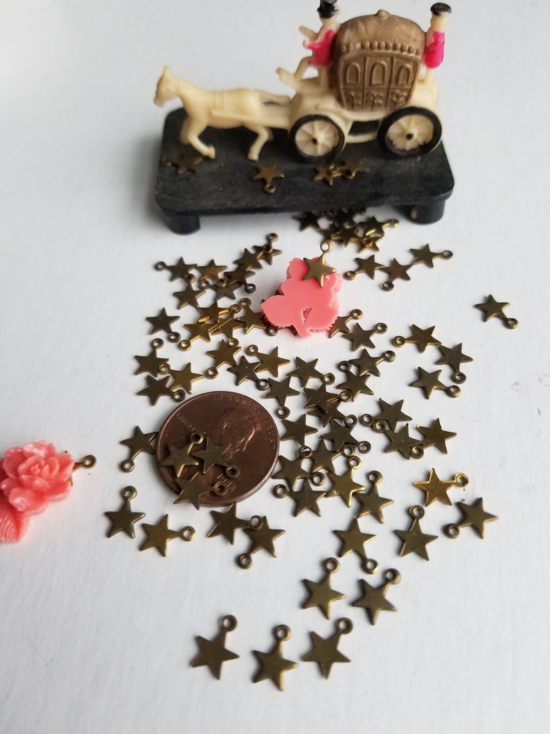 K&ouml;nnte beinhalten: Ein Haufen goldener sternf&ouml;rmiger Charms mit Sprungringen. Die Charms sind auf einer wei&szlig;en Oberfl&auml;che mit einem Penny als Gr&ouml;&szlig;envergleich verstreut. Im Hintergrund befinden sich eine rosafarbene Plastikfigur und eine Spielzeugkutsche.