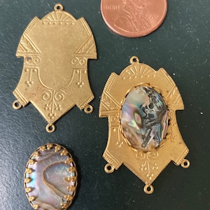 May include: A collection of vintage brass jewelry components, including two ornate pendants and two abalone shell cabochons. A U.S. penny is included for size reference. The components have decorative details and loops for attaching to other pieces.