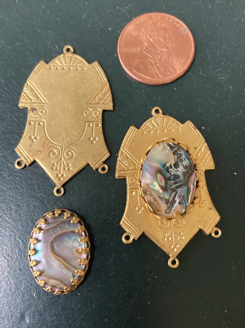 May include: A collection of vintage brass jewelry components, including two ornate pendants and two abalone shell cabochons. A U.S. penny is included for size reference. The components have decorative details and loops for attaching to other pieces.