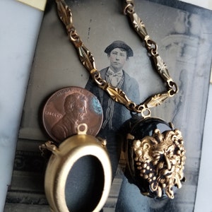 May include: A gold chain necklace with two oval-shaped pendants. One pendant is a blank locket and the other is a black cameo with a gold-toned, detailed, grotesque face.