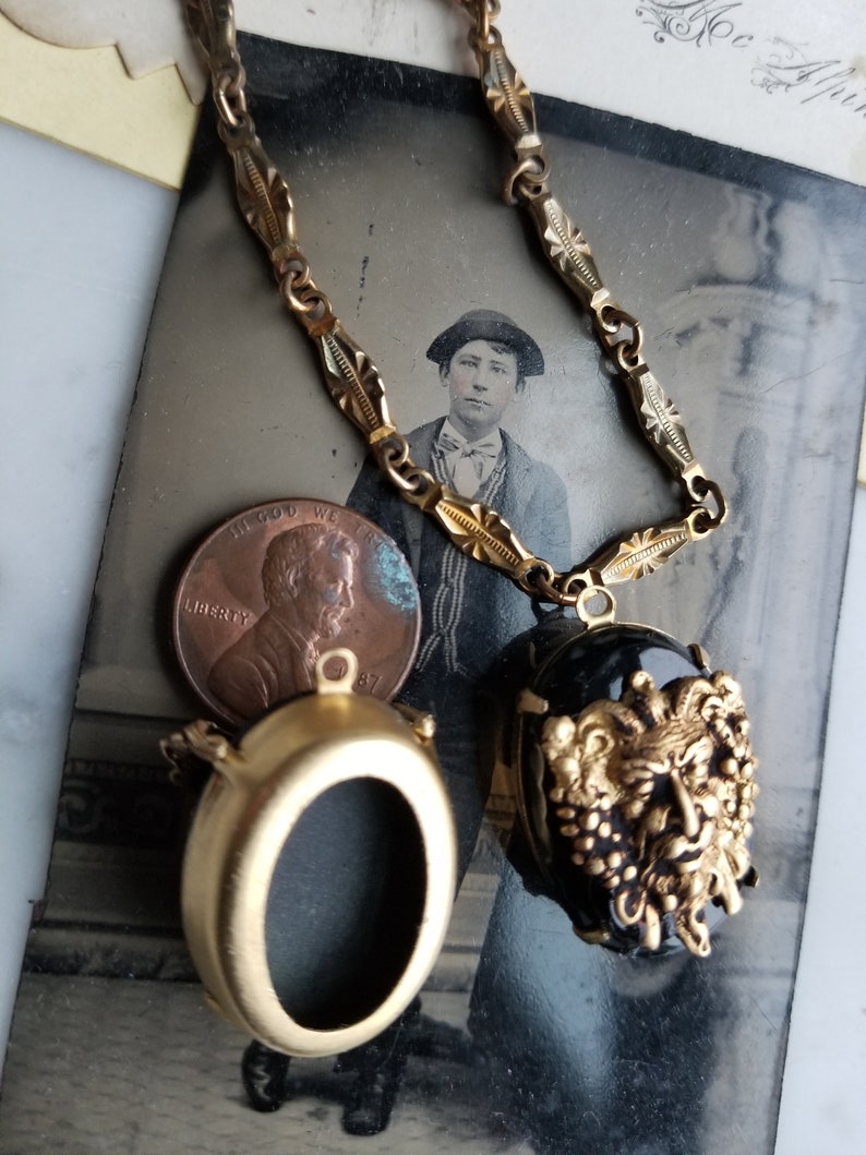 May include: A gold chain necklace with two oval-shaped pendants. One pendant is a blank locket and the other is a black cameo with a gold-toned, detailed, grotesque face.