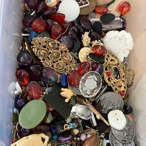 May include: A plastic container filled with a variety of colorful buttons, beads, and jewelry findings. The items include glass, metal, plastic, and wood. There are many different shapes, sizes, and colors.