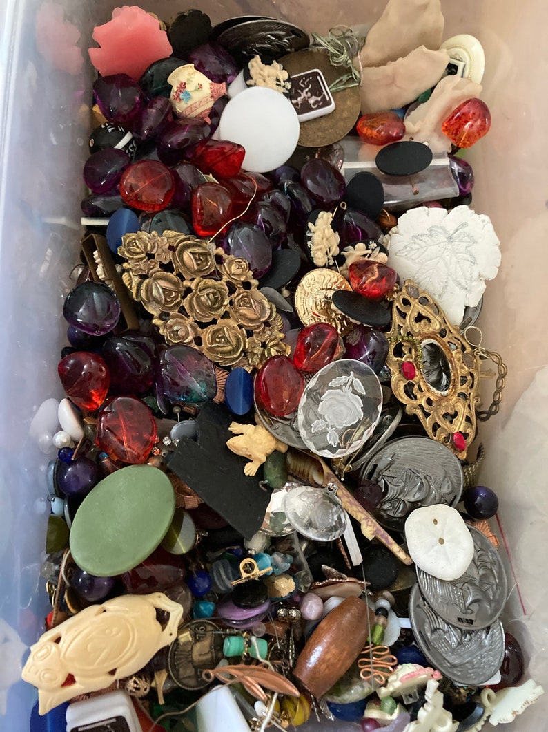 May include: A plastic container filled with a variety of colorful buttons, beads, and jewelry findings. The items include glass, metal, plastic, and wood. There are many different shapes, sizes, and colors.