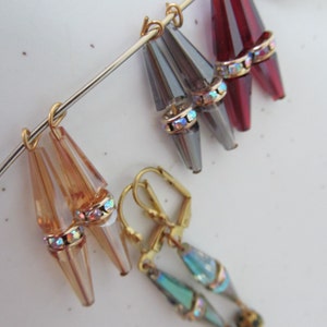 May include: A collection of vintage earrings in various colors. The earrings feature teardrop-shaped, faceted beads in amber, gray, and burgundy, accented with iridescent rhinestones and gold-tone metal. One pair has a teal and gold design.