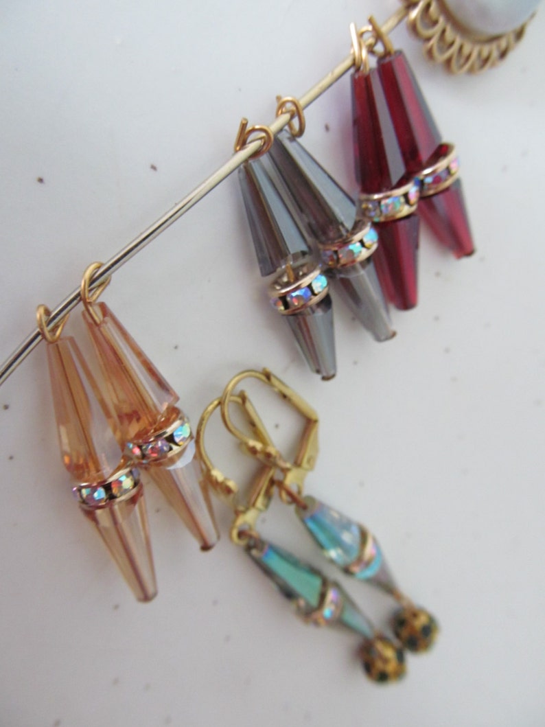 May include: A collection of vintage earrings in various colors. The earrings feature teardrop-shaped, faceted beads in amber, gray, and burgundy, accented with iridescent rhinestones and gold-tone metal. One pair has a teal and gold design.