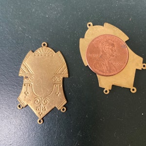 May include: Two antique brass pendants with ornate designs. One pendant is displayed next to a penny for size comparison. The pendants have a shield-like shape with decorative patterns and small holes for attaching to other items. The background is a dark green surface.