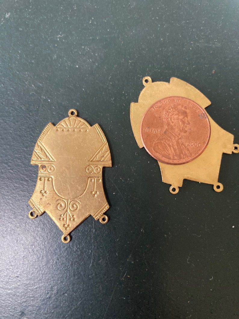 May include: Two antique brass pendants with ornate designs. One pendant is displayed next to a penny for size comparison. The pendants have a shield-like shape with decorative patterns and small holes for attaching to other items. The background is a dark green surface.