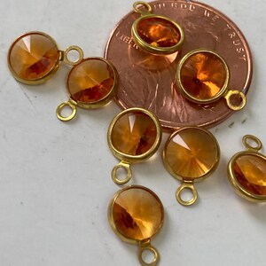 May include: Close-up of several orange-colored, faceted crystal charms with gold-colored metal settings and loops. The charms are round and appear to be new. The background includes a copper-colored coin.