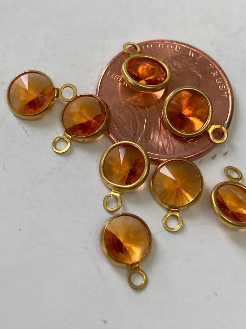 May include: Close-up of several orange-colored, faceted crystal charms with gold-colored metal settings and loops. The charms are round and appear to be new. The background includes a copper-colored coin.