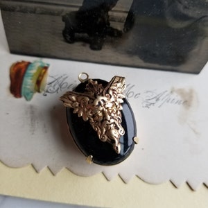 May include: A gold-toned pendant with a black oval cameo featuring a detailed, sculpted, green-faced, bearded, winged creature. The pendant is on a white surface.
