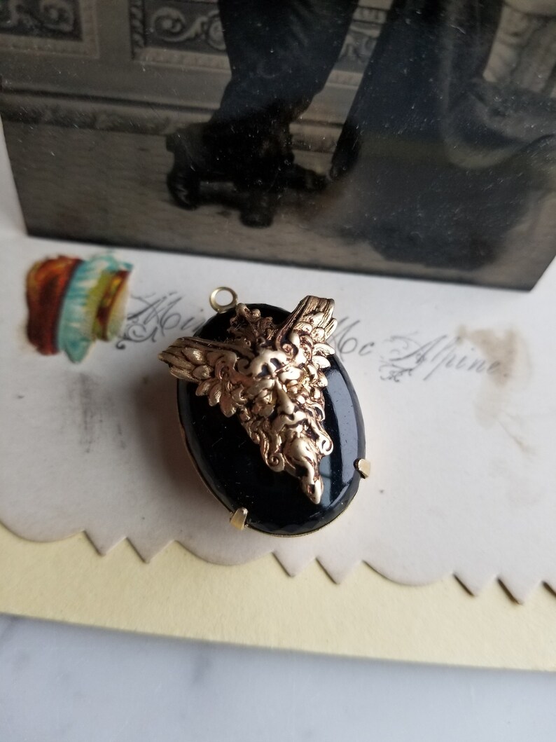 May include: A gold-toned pendant with a black oval cameo featuring a detailed, sculpted, green-faced, bearded, winged creature. The pendant is on a white surface.