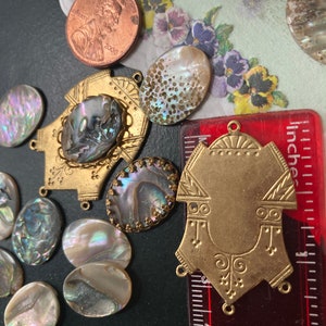 May include: A collection of iridescent abalone shell cabochons and brass findings. Includes a 2000 penny and a ruler showing measurements in inches. The shells vary in shape and size, with a range of colors.