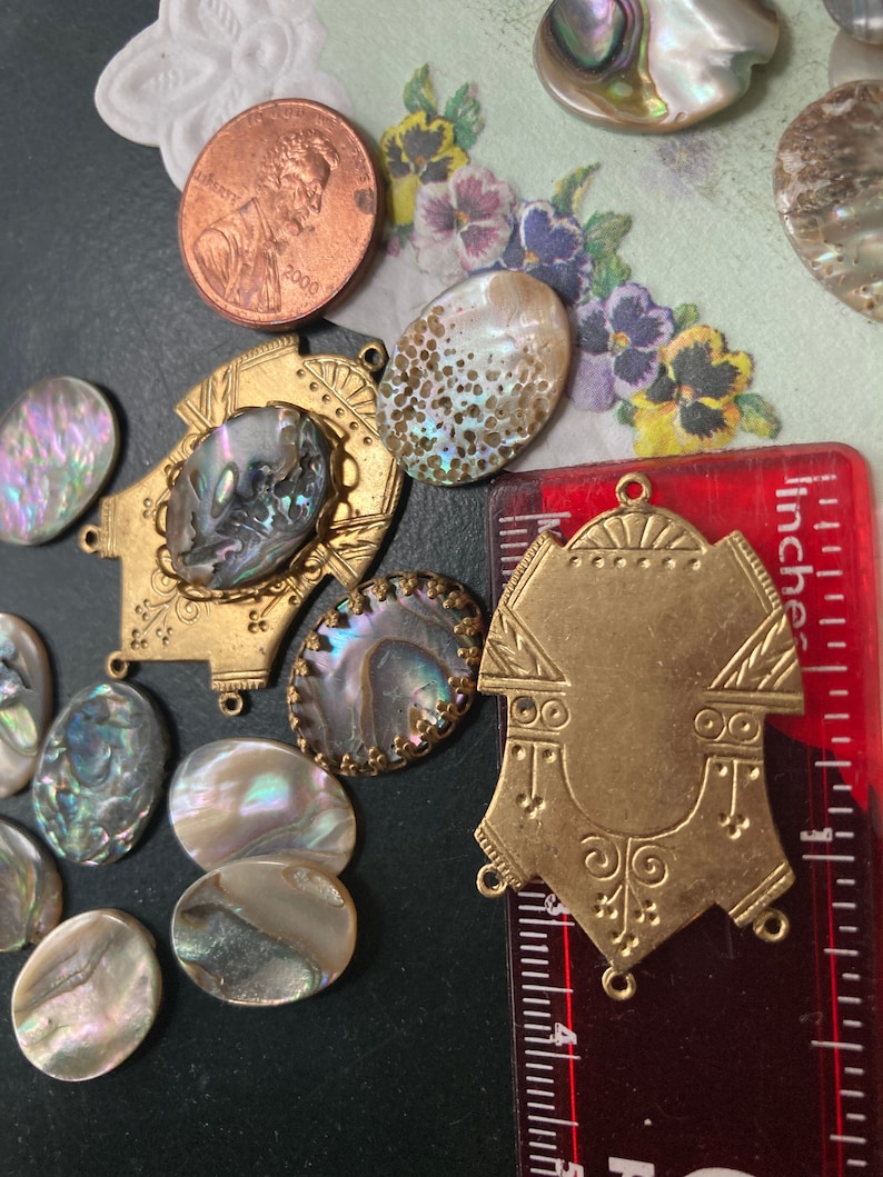 May include: A collection of iridescent abalone shell cabochons and brass findings. Includes a 2000 penny and a ruler showing measurements in inches. The shells vary in shape and size, with a range of colors.
