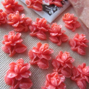 May include: Coral-coloured resin flower embellishments scattered across a neutral surface. Each flower has detailed petals and leaves, suggesting a craft or jewellery-making theme. The flowers are approximately 2.5 cm in diameter.