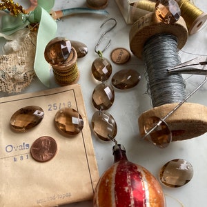 May include: A collection of vintage glass beads in shades of brown, with a spool of wire, a spool of gold thread, and a vintage ornament. The beads are oval shaped and have a faceted design. The ornament is red and gold with a striped pattern. The image also includes a vintage spool of thread with a label that reads "Vintage".