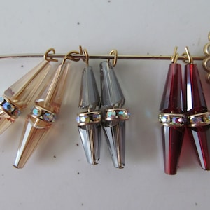 May include: Three pairs of crystal teardrop earrings with gold-toned metal findings. The earrings are in shades of beige, grey, and red.