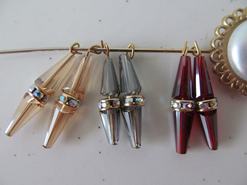 May include: Three pairs of crystal teardrop earrings with gold-toned metal findings. The earrings are in shades of beige, grey, and red.