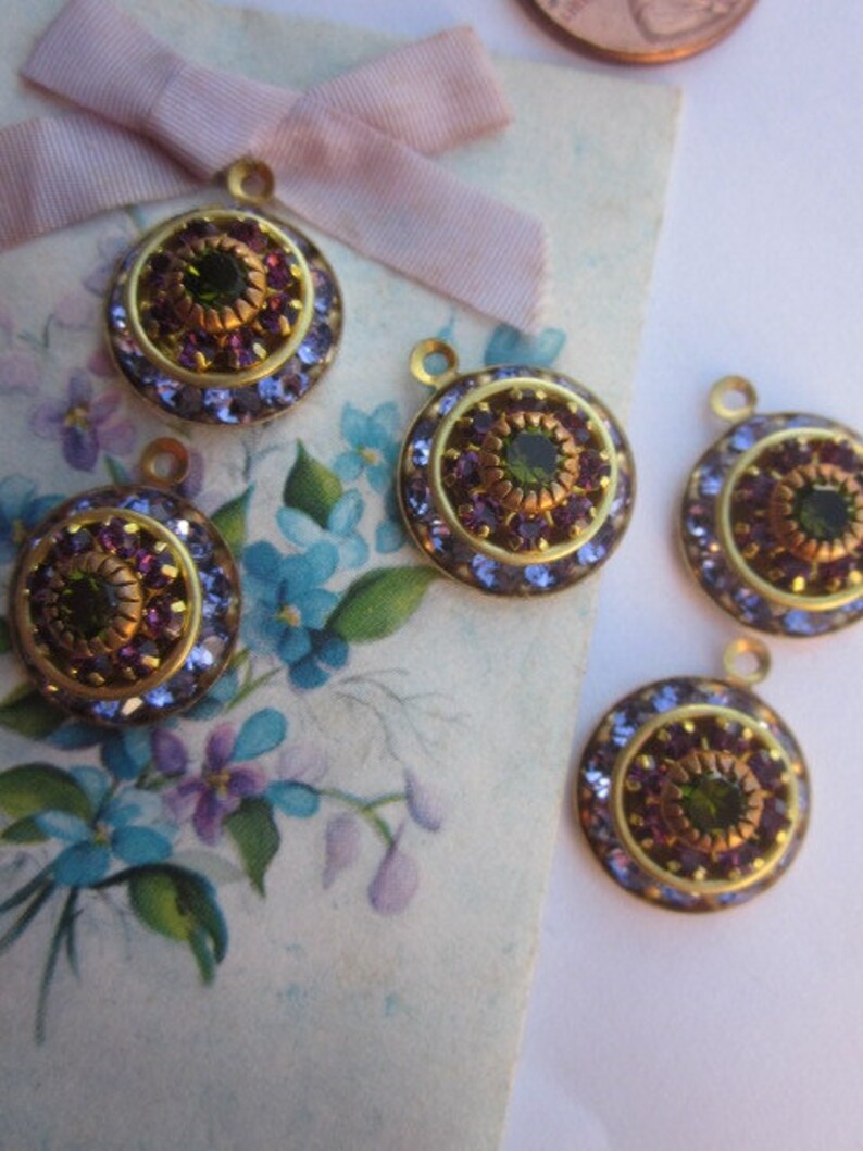 May include: Five gold-toned round pendants with a green and purple rhinestone design. Each pendant has a loop at the top for attaching to a chain or other jewelry.