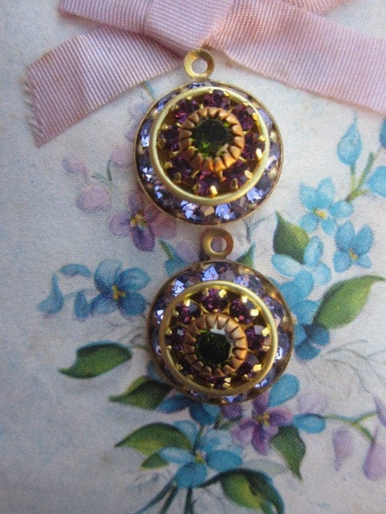 May include: Two gold-toned round metal embellishments with a center design of a green jewel surrounded by purple jewels. The embellishments are on a floral fabric background.