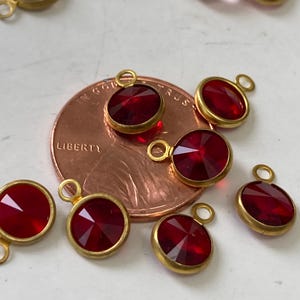 May include: Close-up of several red gemstone pendants with gold-colored bezels. The pendants are round and oval-shaped, with a faceted cut that catches the light. A penny is in the background, showing the word "LIBERTY".