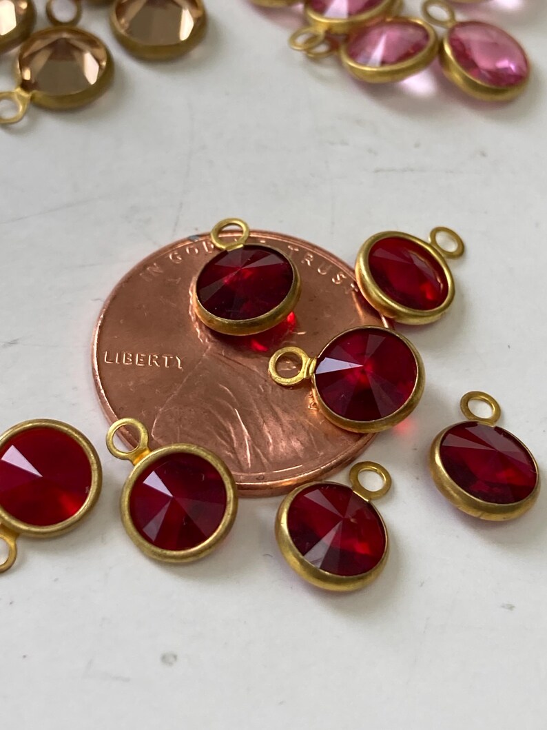 May include: Close-up of several red gemstone pendants with gold-colored bezels. The pendants are round and oval-shaped, with a faceted cut that catches the light. A penny is in the background, showing the word "LIBERTY".