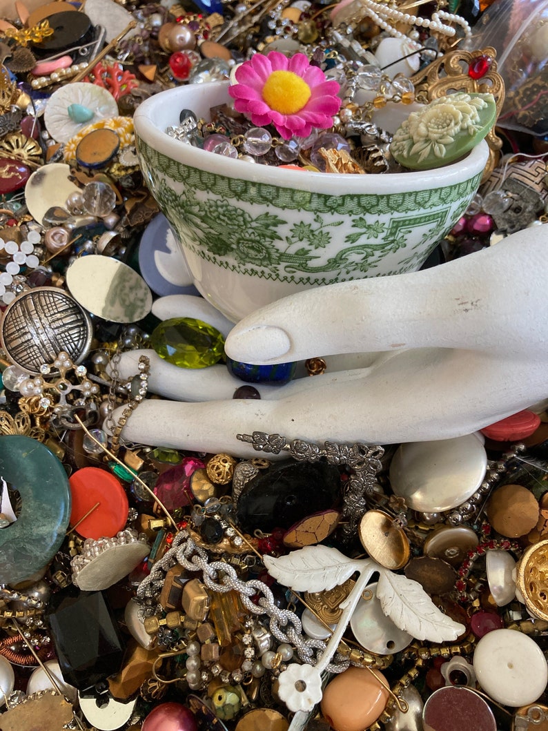 May include: A white ceramic teacup with a green floral pattern filled with colorful beads, buttons, and jewelry. A pink flower with a yellow center sits on top of the beads.
