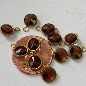 May include: Close-up of several small, round, faceted, brown gemstone charms with gold-colored metal settings and loops. A penny is included for size reference. The charms are likely for jewelry making.