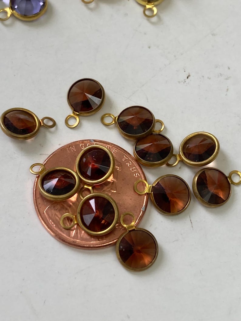 May include: Close-up of several small, round, faceted, brown gemstone charms with gold-colored metal settings and loops. A penny is included for size reference. The charms are likely for jewelry making.