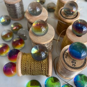 Peut inclure: Assortiment de billes de verre iris&eacute;es sur des bobines de fil. Les billes sont de diff&eacute;rentes nuances de bleu, vert et violet. Les bobines sont en bois et ont une finition dor&eacute;e ou argent&eacute;e.