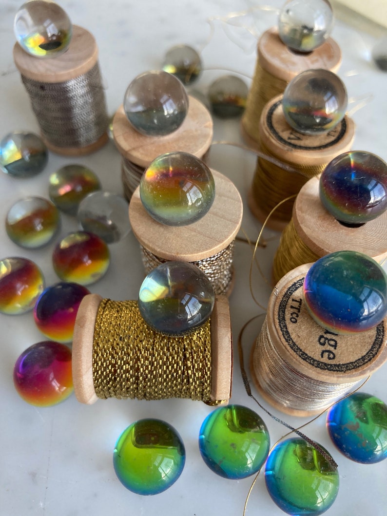 Peut inclure: Assortiment de billes de verre iris&eacute;es sur des bobines de fil. Les billes sont de diff&eacute;rentes nuances de bleu, vert et violet. Les bobines sont en bois et ont une finition dor&eacute;e ou argent&eacute;e.