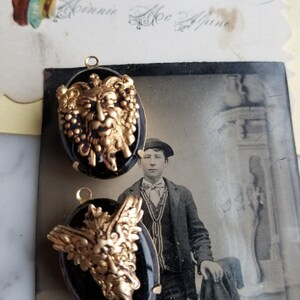 May include: Two black and gold oval-shaped pendants with a detailed design of a face with a crown and vines. The pendants are hanging from a white surface.
