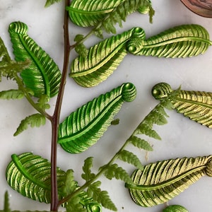May include: A collection of green and gold leaf-shaped metal embellishments. The leaves have a textured surface and are arranged on a white background with a fern branch.