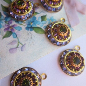 May include: Four gold-toned round pendants with a central green gemstone surrounded by smaller purple gemstones. Each pendant has a loop for attaching to a chain or other jewelry.