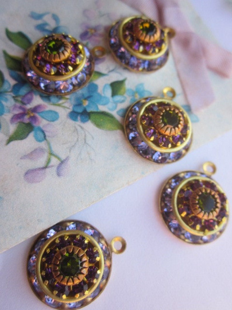 May include: Four gold-toned round pendants with a central green gemstone surrounded by smaller purple gemstones. Each pendant has a loop for attaching to a chain or other jewelry.