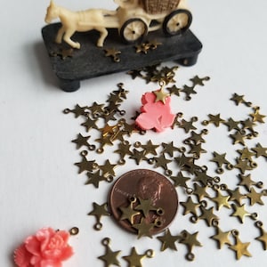 K&ouml;nnte beinhalten: Ein Haufen goldener sternf&ouml;rmiger Charms mit Sprungringen. Die Charms sind auf einer wei&szlig;en Oberfl&auml;che mit einem Penny und zwei rosafarbenen Plastik-Rosen-Charms verstreut.