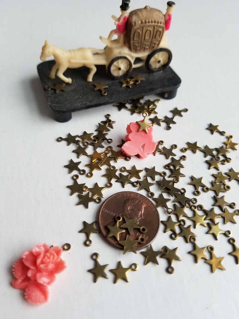 K&ouml;nnte beinhalten: Ein Haufen goldener sternf&ouml;rmiger Charms mit Sprungringen. Die Charms sind auf einer wei&szlig;en Oberfl&auml;che mit einem Penny und zwei rosafarbenen Plastik-Rosen-Charms verstreut.
