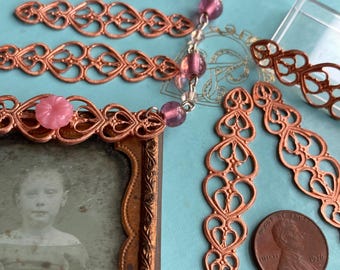 Può includere: Una collana color rame con un ciondolo che presenta una fotografia vintage. La collana ha una catena con perline di vetro rosa e più ciondoli a forma di cuore in filigrana di rame. Il ciondolo ha una cornice decorativa e un accento di fiore rosa.