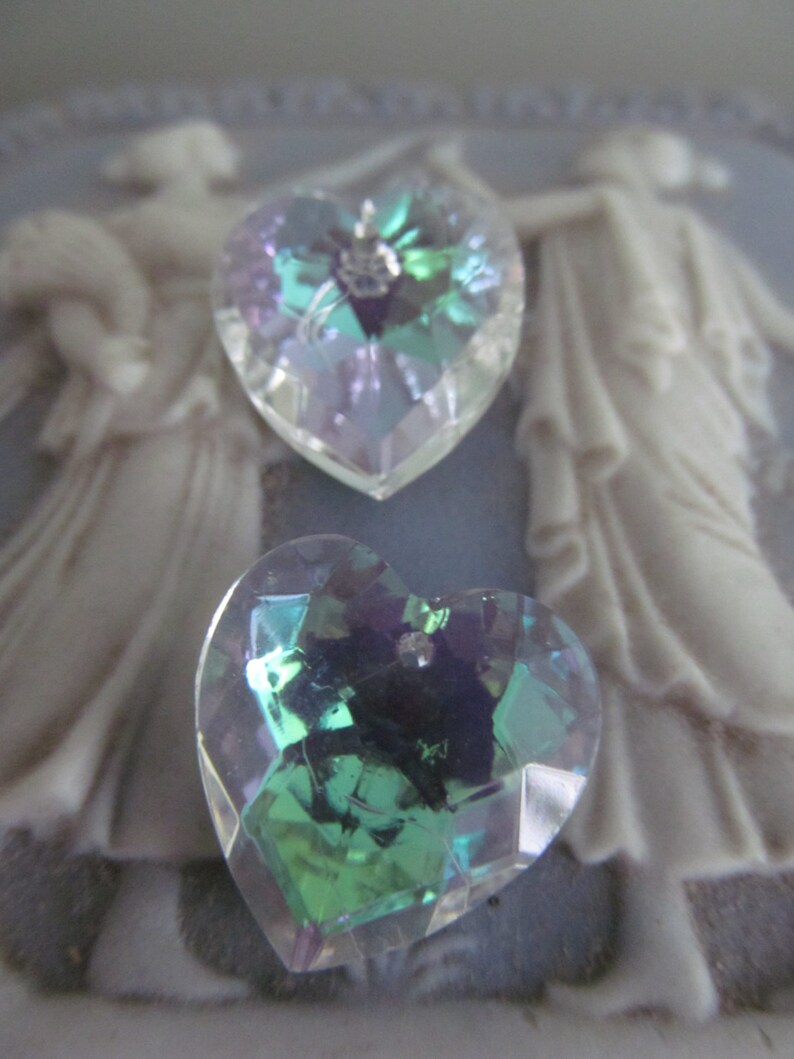 May include: Two iridescent heart-shaped crystal charms. One charm is a light blue and purple color, the other is a dark green and purple color.
