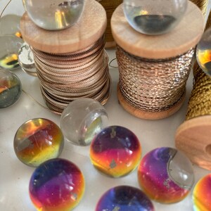 Peut inclure: Une collection de billes en verre de diff&eacute;rentes couleurs, dont du transparent, de l'iridescent et de l'or. Les billes sont dispos&eacute;es sur une surface blanche avec des bobines de fil d'or et d'argent.