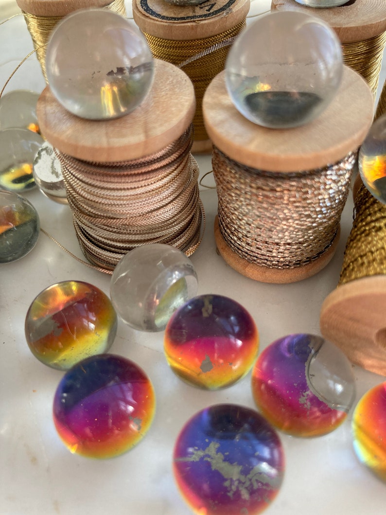Peut inclure: Une collection de billes en verre de diff&eacute;rentes couleurs, dont du transparent, de l'iridescent et de l'or. Les billes sont dispos&eacute;es sur une surface blanche avec des bobines de fil d'or et d'argent.