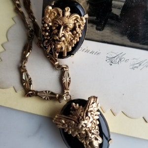 May include: A gold chain necklace with two black cameo pendants. The cameos feature gold-toned, detailed designs of a face with foliage.