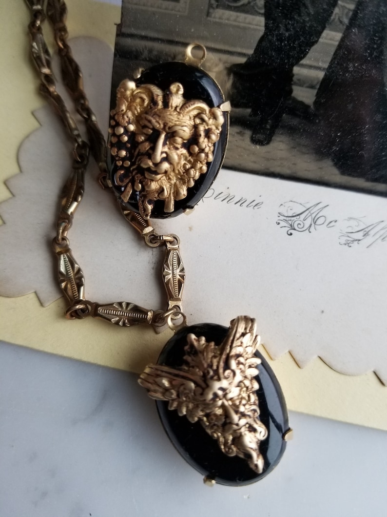 May include: A gold chain necklace with two black cameo pendants. The cameos feature gold-toned, detailed designs of a face with foliage.