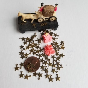 K&ouml;nnte beinhalten: Ein Haufen goldfarbener sternf&ouml;rmiger Charms mit Sprungringen, ein rosafarbener Blumencharm und eine Miniaturkutsche mit zwei Figuren auf einem schwarzen Sockel. Ein Cent-St&uuml;ck ist zur Gr&ouml;&szlig;enbestimmung enthalten.