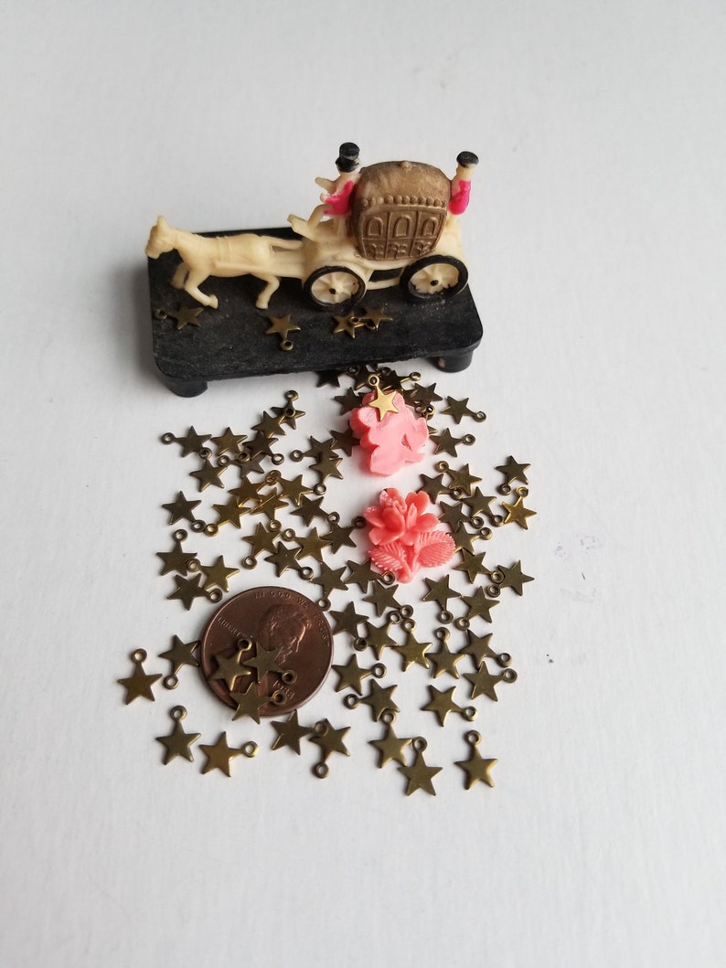 K&ouml;nnte beinhalten: Ein Haufen goldfarbener sternf&ouml;rmiger Charms mit Sprungringen, ein rosafarbener Blumencharm und eine Miniaturkutsche mit zwei Figuren auf einem schwarzen Sockel. Ein Cent-St&uuml;ck ist zur Gr&ouml;&szlig;enbestimmung enthalten.
