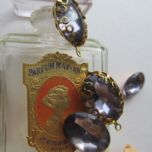 May include: A vintage glass bottle with a gold label that reads "PARFUM MARIANA" and "JN. GIRAUD PARIS". The bottle is decorated with three gold-toned metal embellishments with dark grey glass stones.